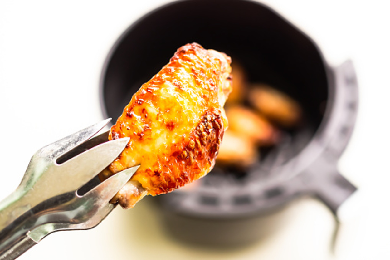 A chicken piece held by a tong and an air fryer against white background
