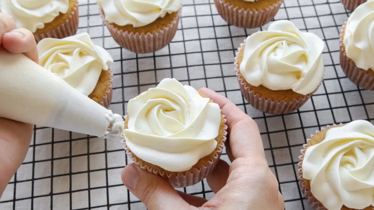 Human hands piping rose flower frosting on vanilla cupcakes.
