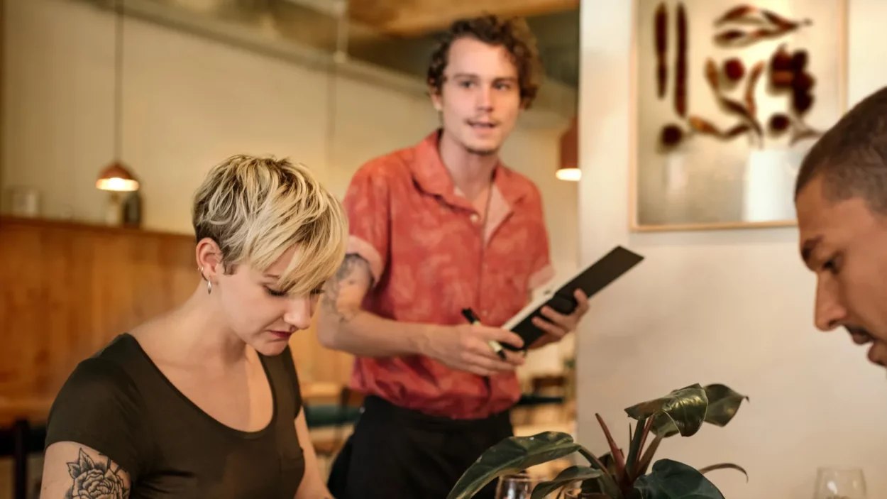 Diverse group of young friends ordering food from a waiter while sitting together at a table.