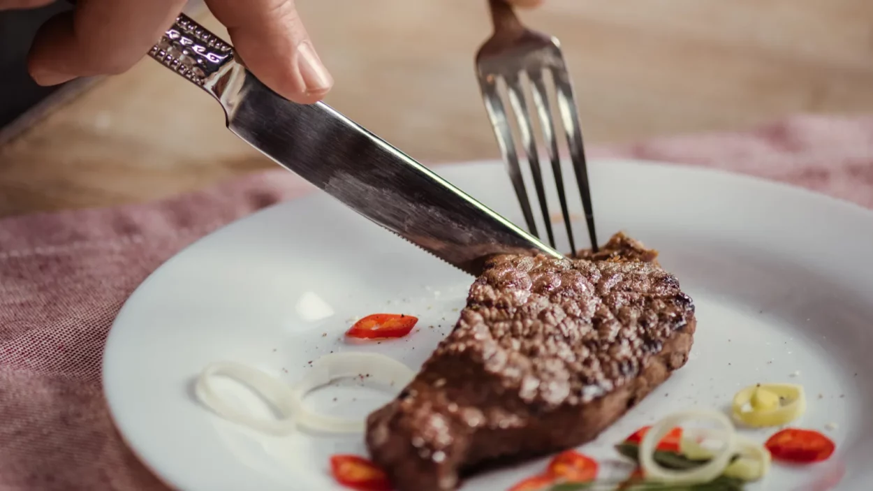 Close-up partial view of man eating steak with fork and knife.