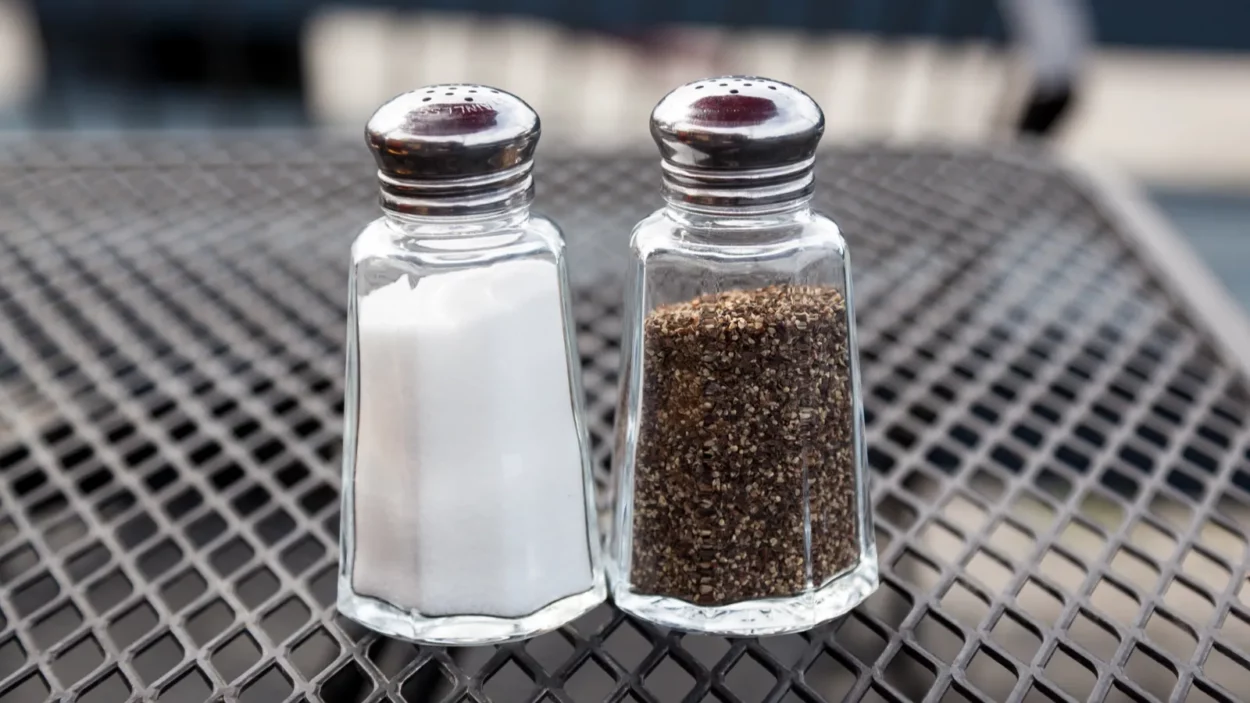 Classic glass salt and pepper shakers on a metal grate table at a patio of a restaurant.