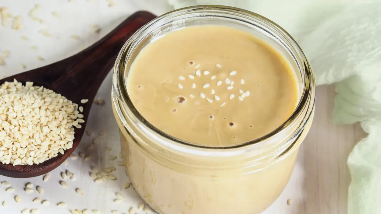 Top view of mason jar filled with tahini. Wooden spoon overflowing with sesame seeds.