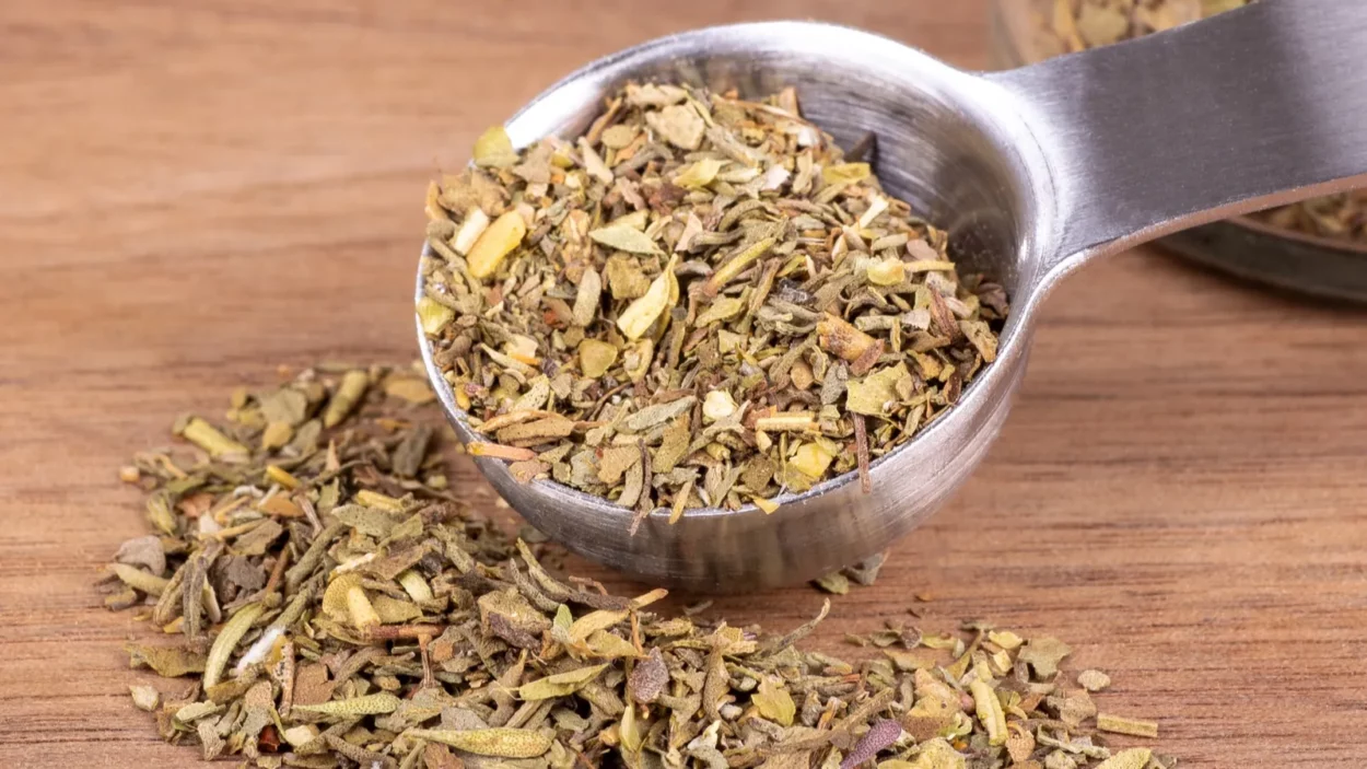 Closeup of chopped Italian seasoning and a measuring spoon on a wooden table.