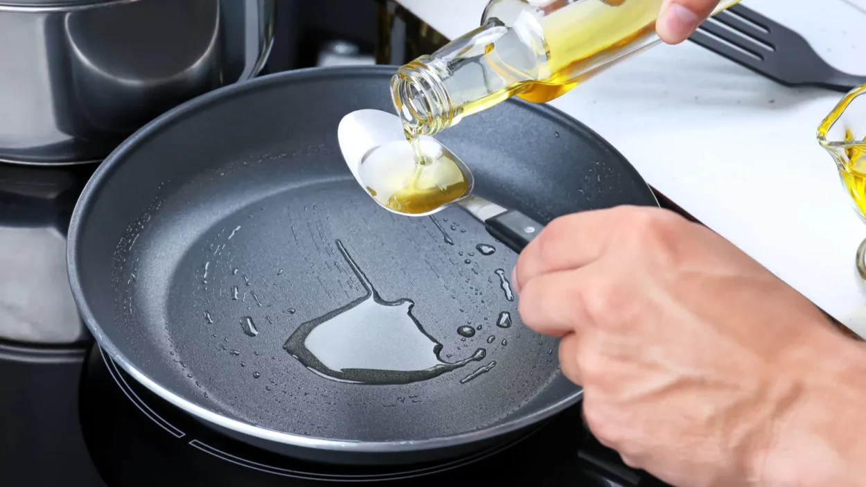 Man pouring cooking oil from bottle into spoon over frying pan in kitchen.