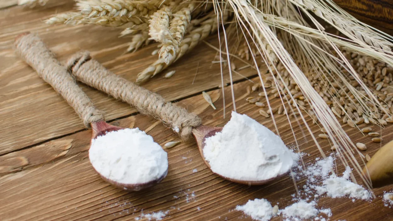 On a wooden table lie ears of wheat and two tablespoons of flour scattered on the table.